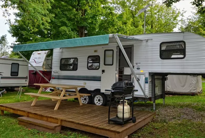 Caravane avec terrasse en bois, barbecue visible.